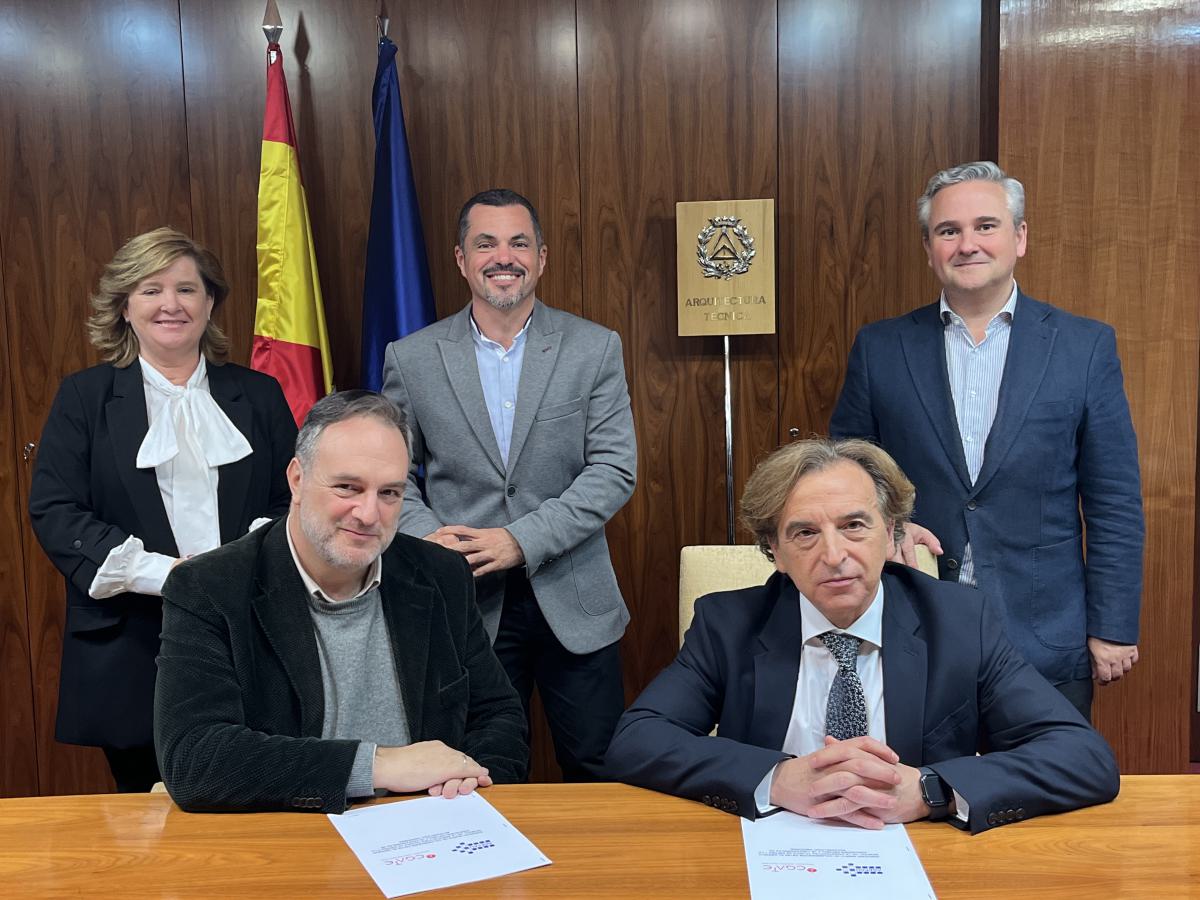 cinco personas, dos personas delante sentadas y tres detrás en pie. En la mesa dos ejemplares de un documento. Al fondo bandera de España