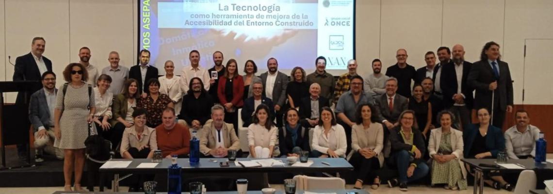 Foto de grupo de los participantes, en varias filas, con la pantalla detrás con el título del evento